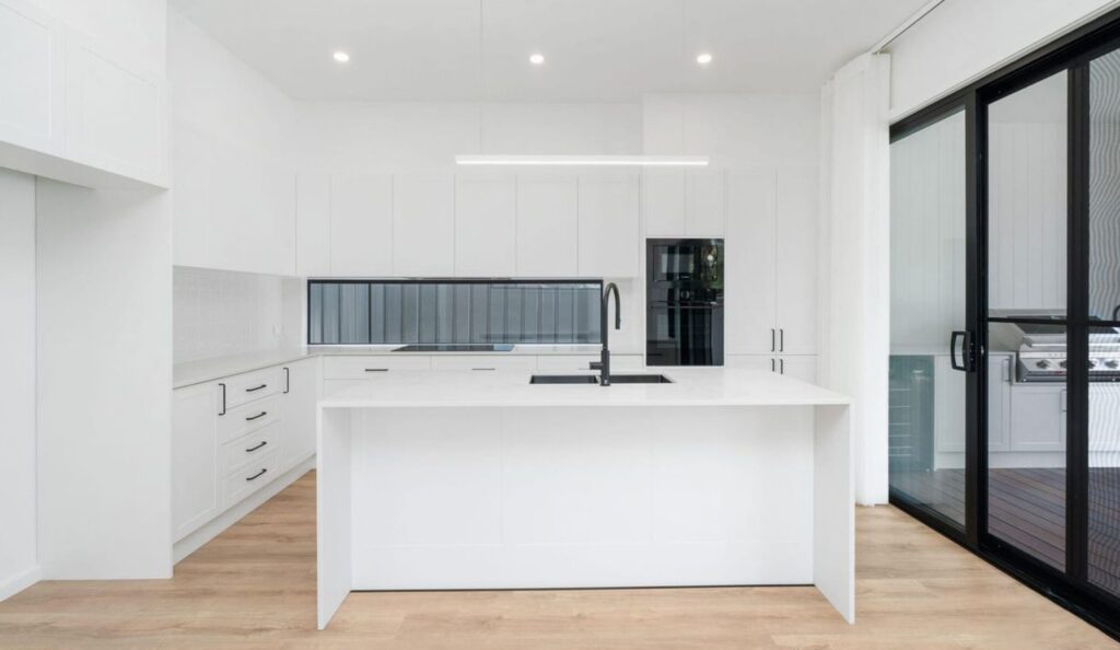 A bright, modern white kitchen featuring a prominent central waterfall island with a black sink and faucet.